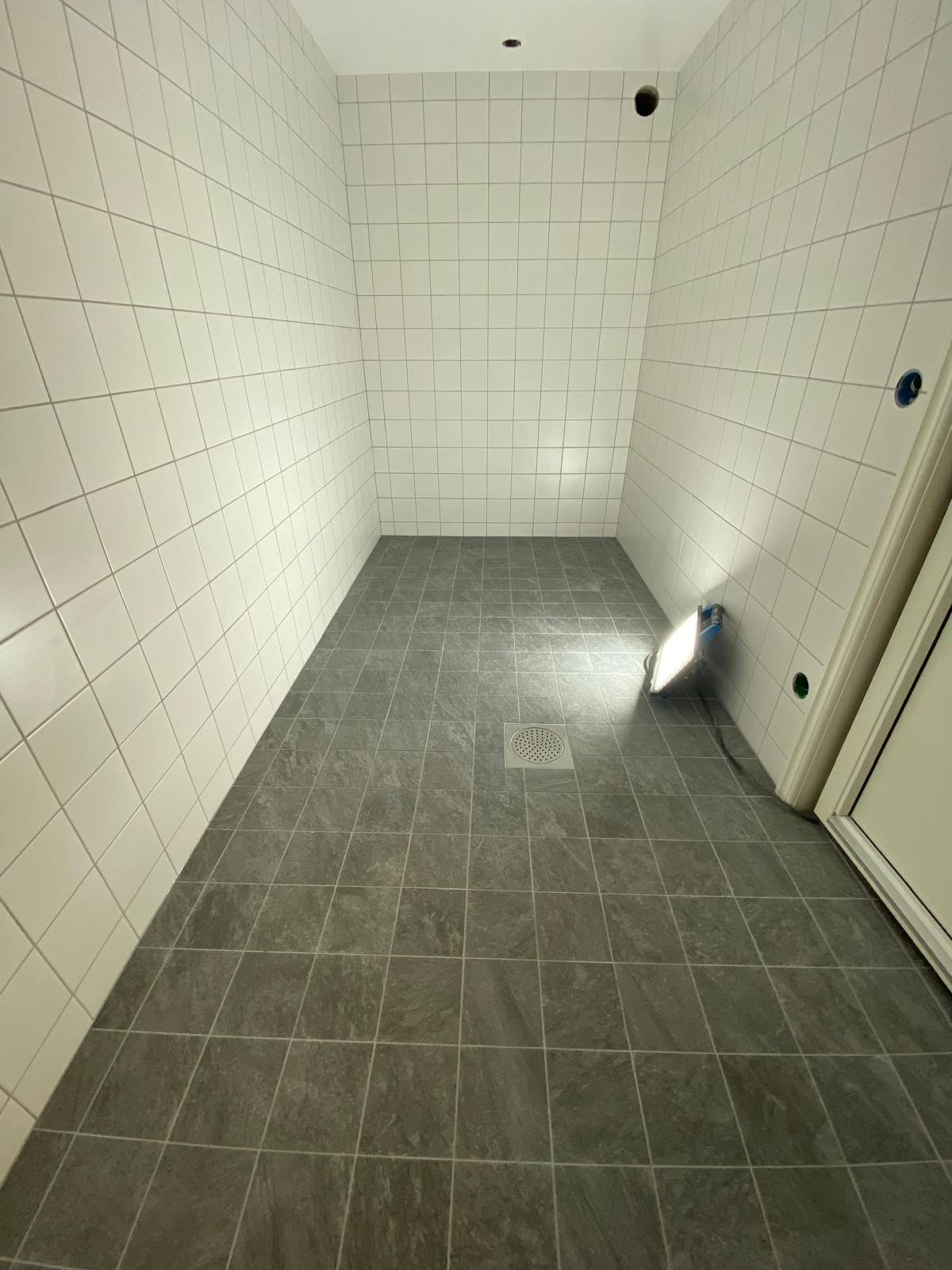 Källarutrymme under renovering med vitt kakel, grå klinker och golvbrunn.