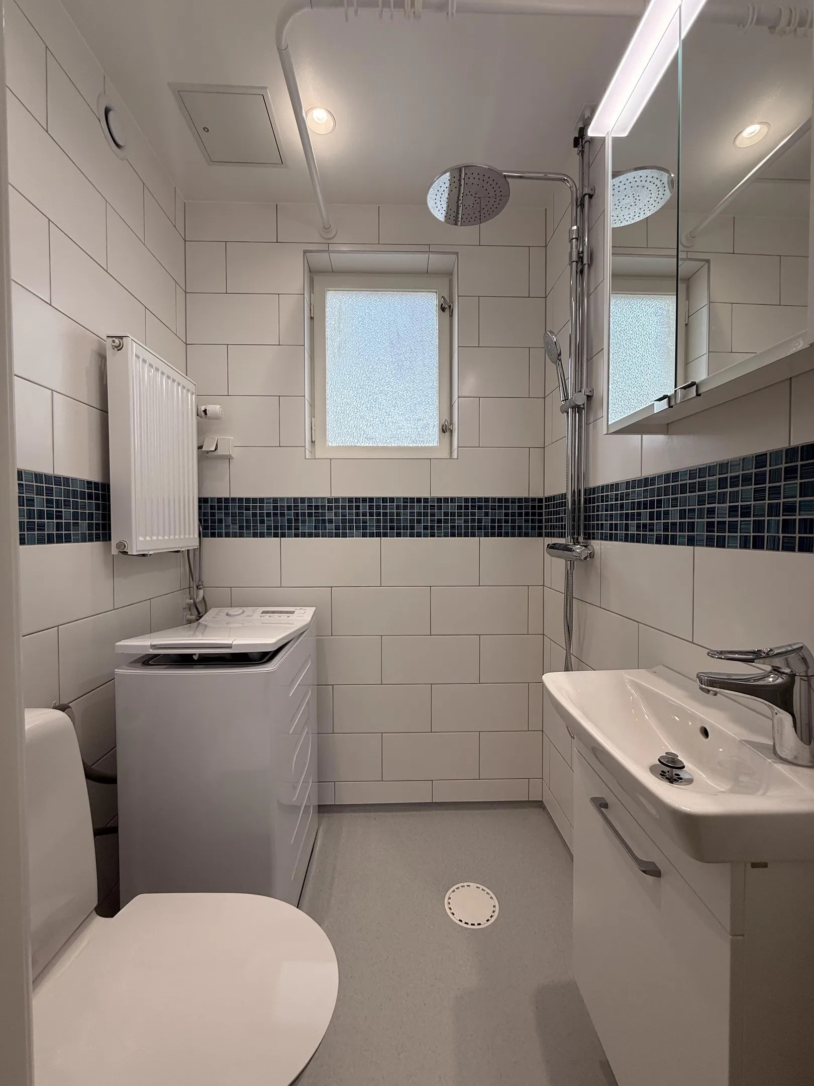 Renoverat litet badrum med vitt kakel, blå mosaik, takdusch och tvättmaskin.